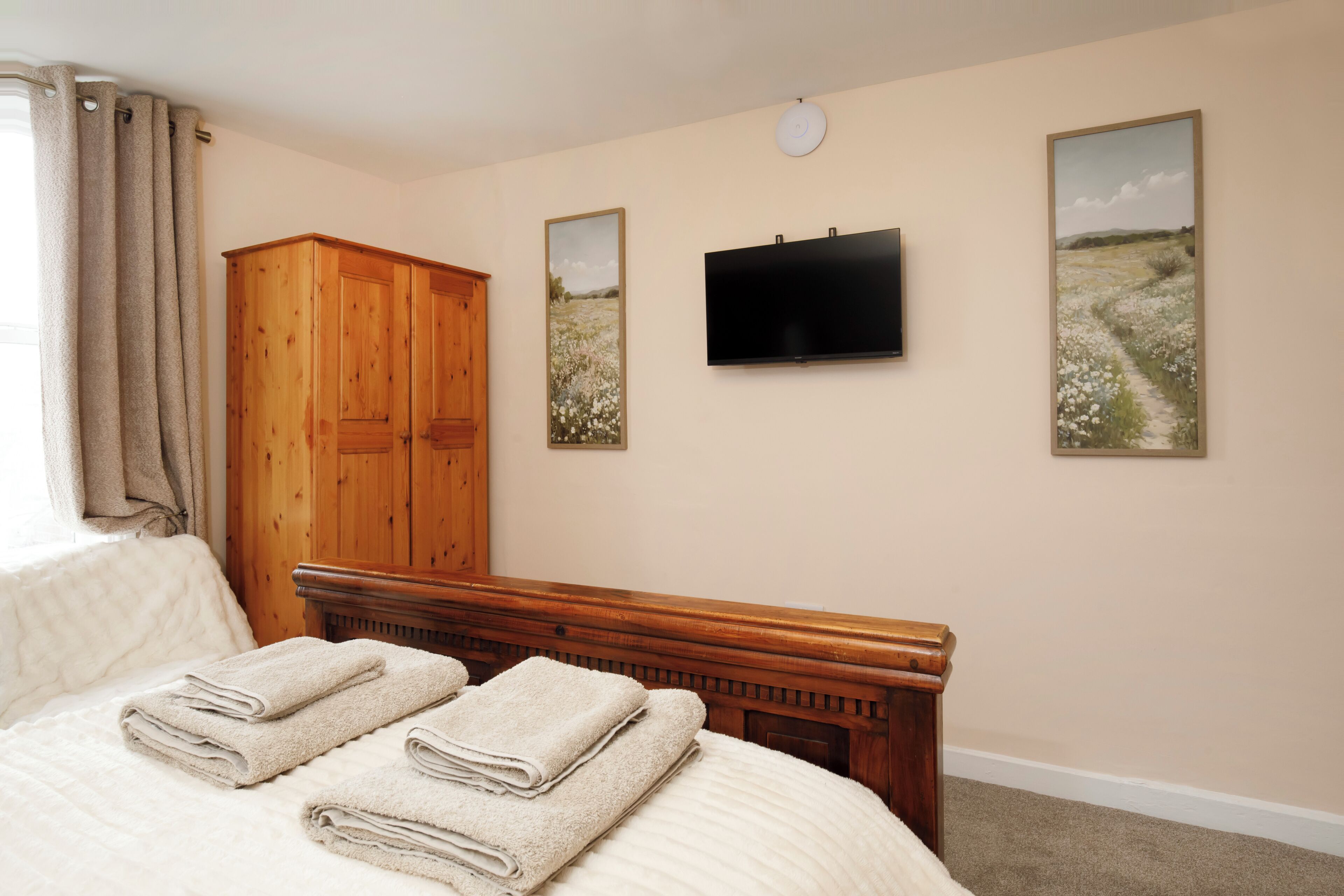 A bedroom with a bed, a wooden wardrobe, a flat-screen TV, and two framed pictures on the wall.