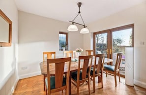 Une salle à manger avec une table et des chaises en bois, un lustre et une vue sur l’extérieur, visible à travers une porte vitrée.