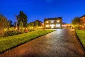 A two-story house with a driveway leading to the front door, surrounded by well-lit trees and a lawn. - CH-Stylish Home with Hot Tub & Garden (West Yorkshire)
