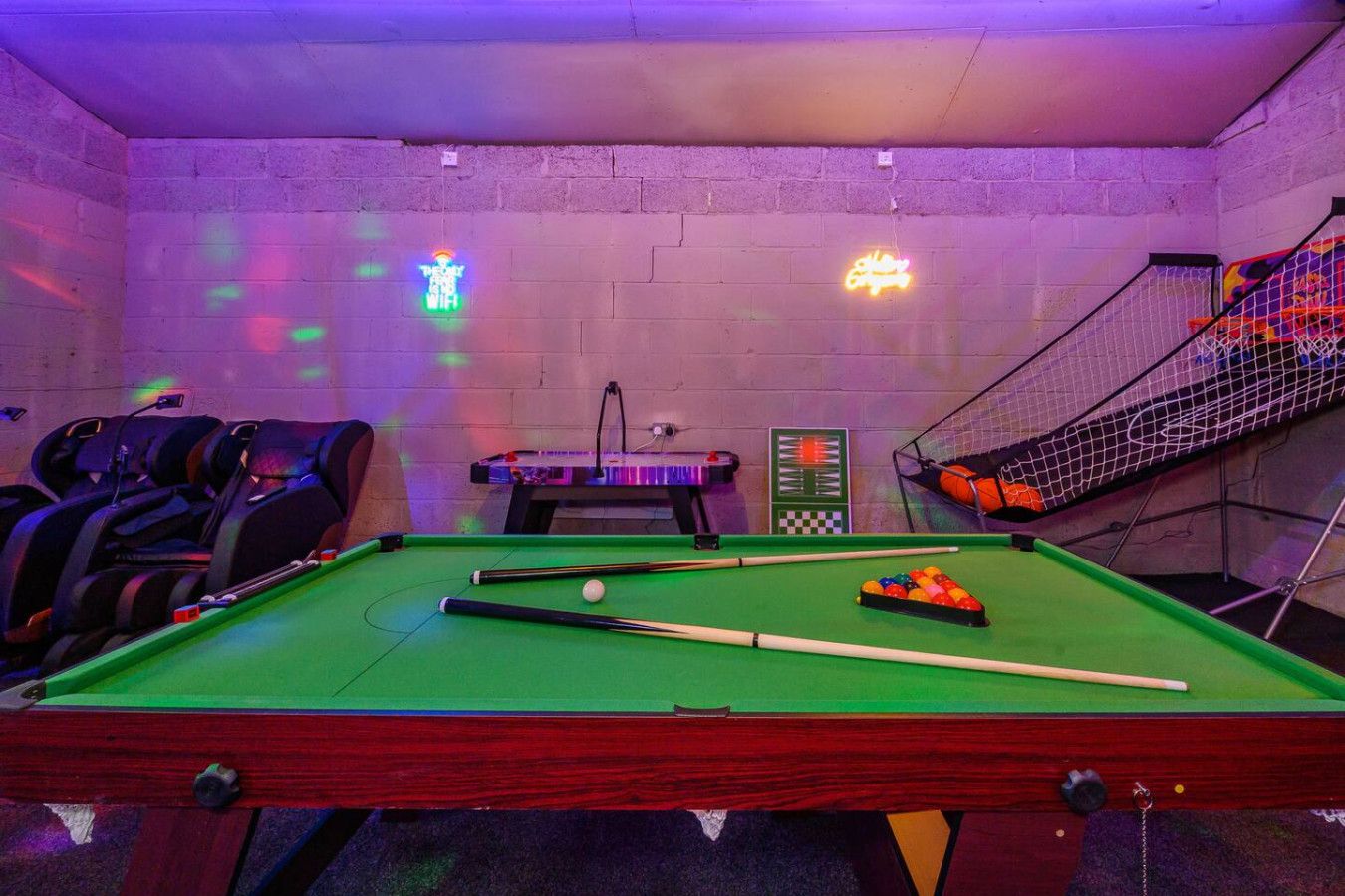 A pool table with balls, a billiard cue, and a rack of billiard balls. There are chairs and a small table with a neon sign.