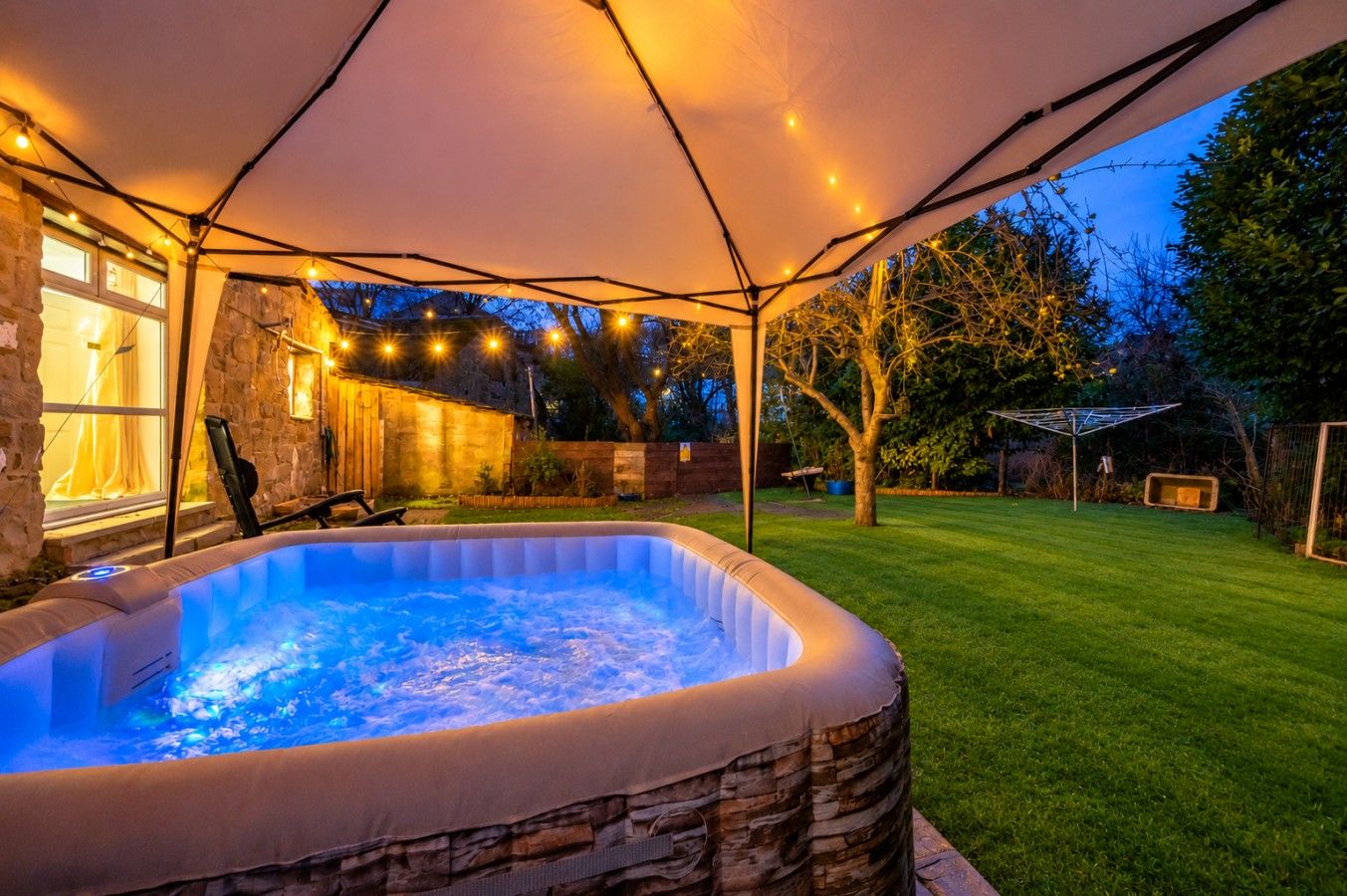 A hot tub under a covered area with string lights, a stone wall, and a lawn.