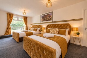 A hotel room with two beds, a large window with curtains, a wall-mounted picture, and a small table with a lamp and plant. - Cozy Farm house with Hot Tub retreat (West Yorkshire)