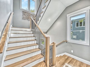 Eine Treppe mit Holzstufen und Metallgeländern, ein Fenster mit Blick auf ein Haus und eine Wandleuchte.