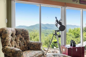 A room with a large window, a telescope, a floral armchair, and a red suitcase.