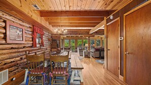 A log cabin interior with a dining area, wooden walls, and a view through large windows.