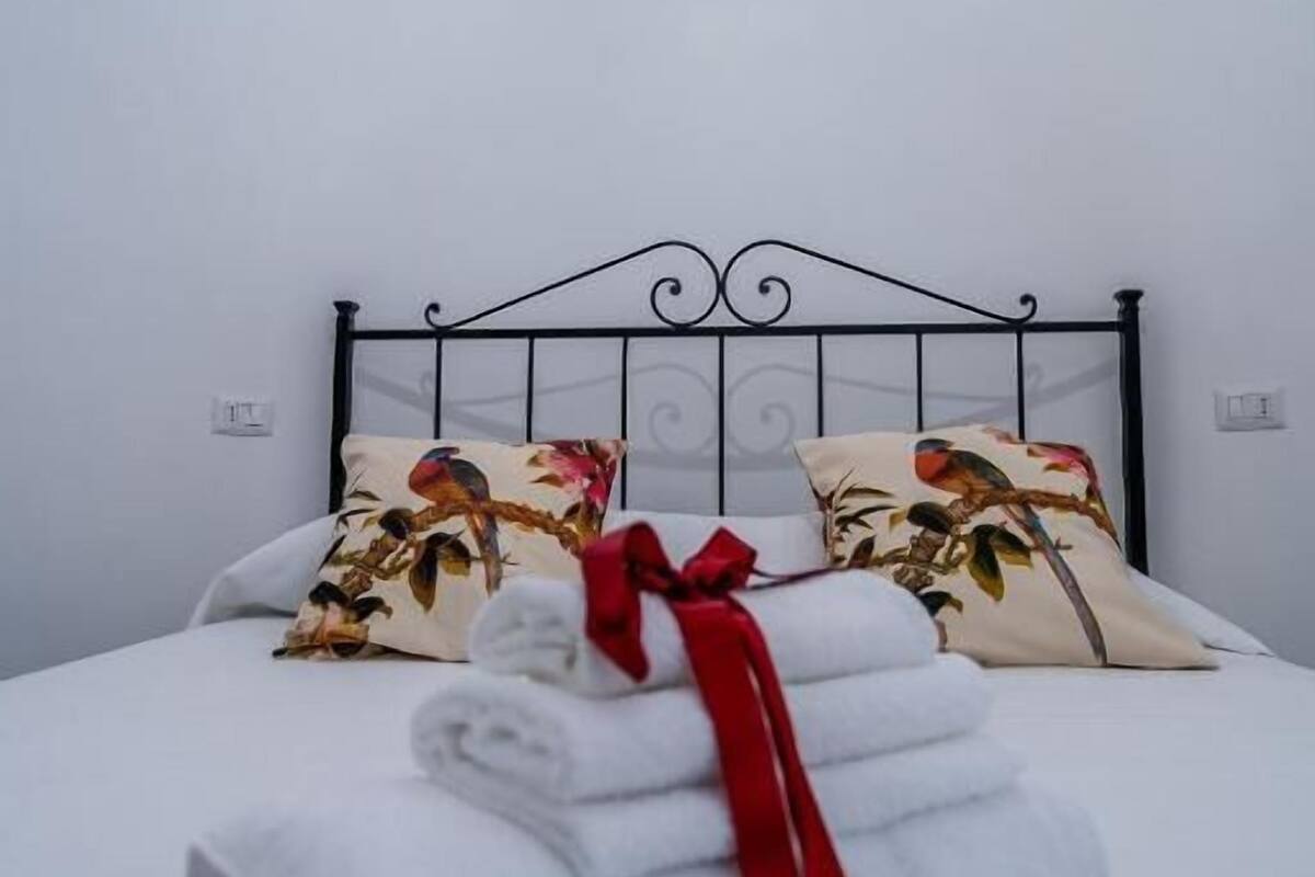Una cama bien tendida con sábanas blancas y un lazo rojo, una ventana gótica arriba y almohadones florales.