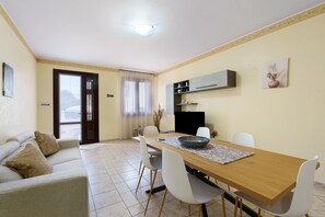 Un espace repas comprenant une table en bois, des chaises blanches, un canapé avec des coussins, un coin cuisine avec des placards et une porte donnant sur l’extérieur.
