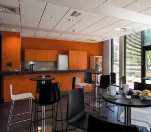 A modern kitchen with orange cabinets, a refrigerator, and a coffee machine. There are black chairs and a round table with a white mug and a plate of food. - Adonis Dijon Double Apartment (Dijon)