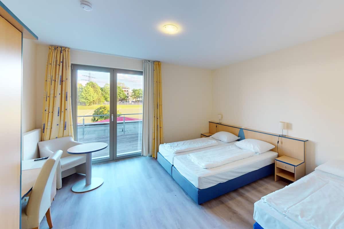 Habitación de hotel con cama, escritorio, silla y vista a un campo deportivo por una puerta corrediza.
