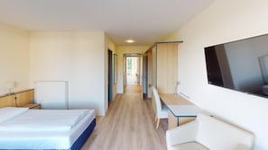 Habitación de hotel con cama, escritorio, silla, televisor y vista al pasillo.