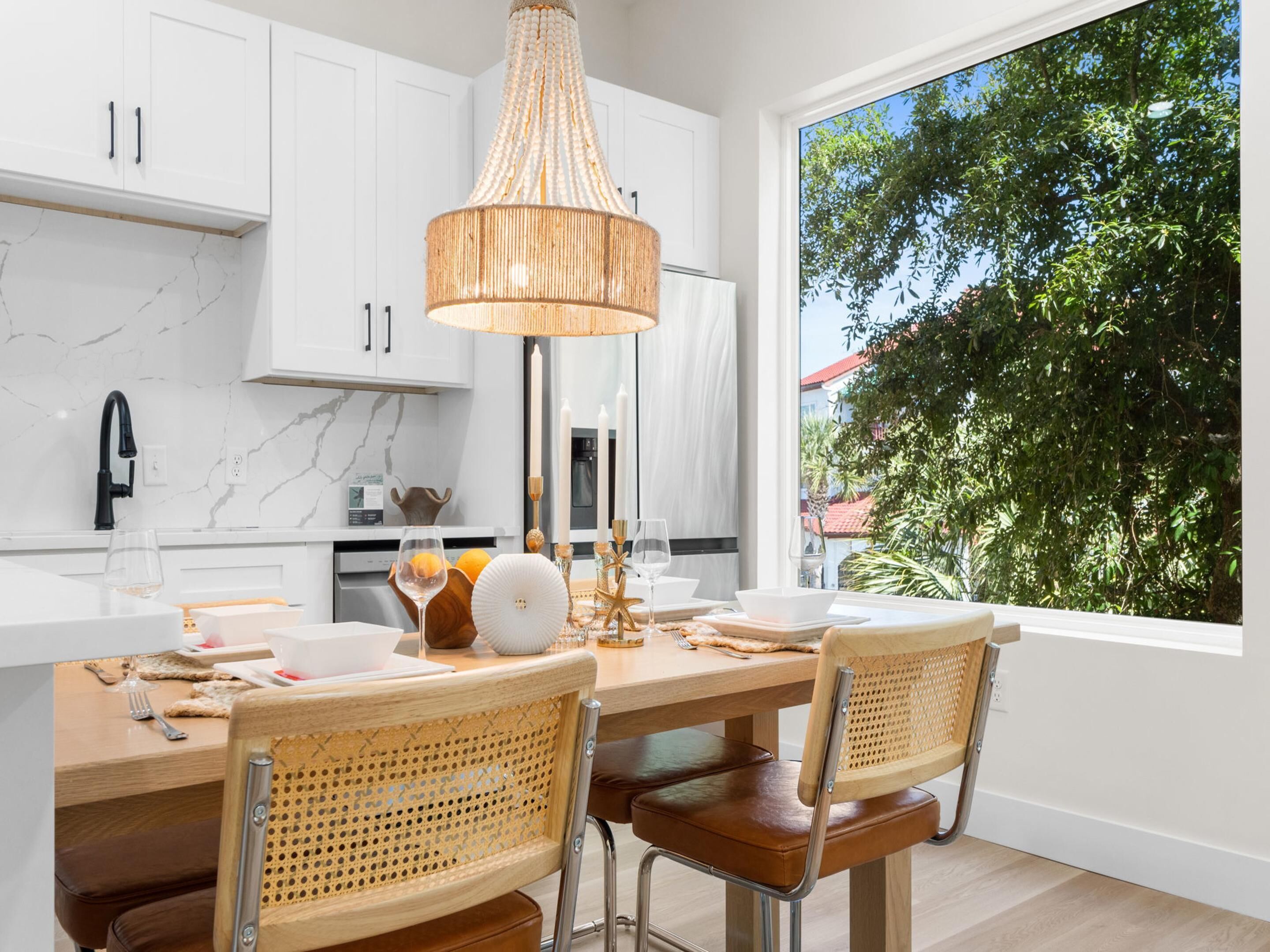 Une cuisine moderne avec des meubles blancs, un pare-feu en marbre, une grande fenêtre donnant sur un espace verdoyant et un coin repas avec des chaises en osier et un îlot central.