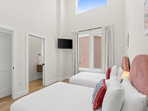 A bedroom with two beds, a TV, a window with curtains, and a bathroom visible through an open door. - 3014 Coral Blossom A | 0.3mi to Beach, Game Room (Panama City Beach)