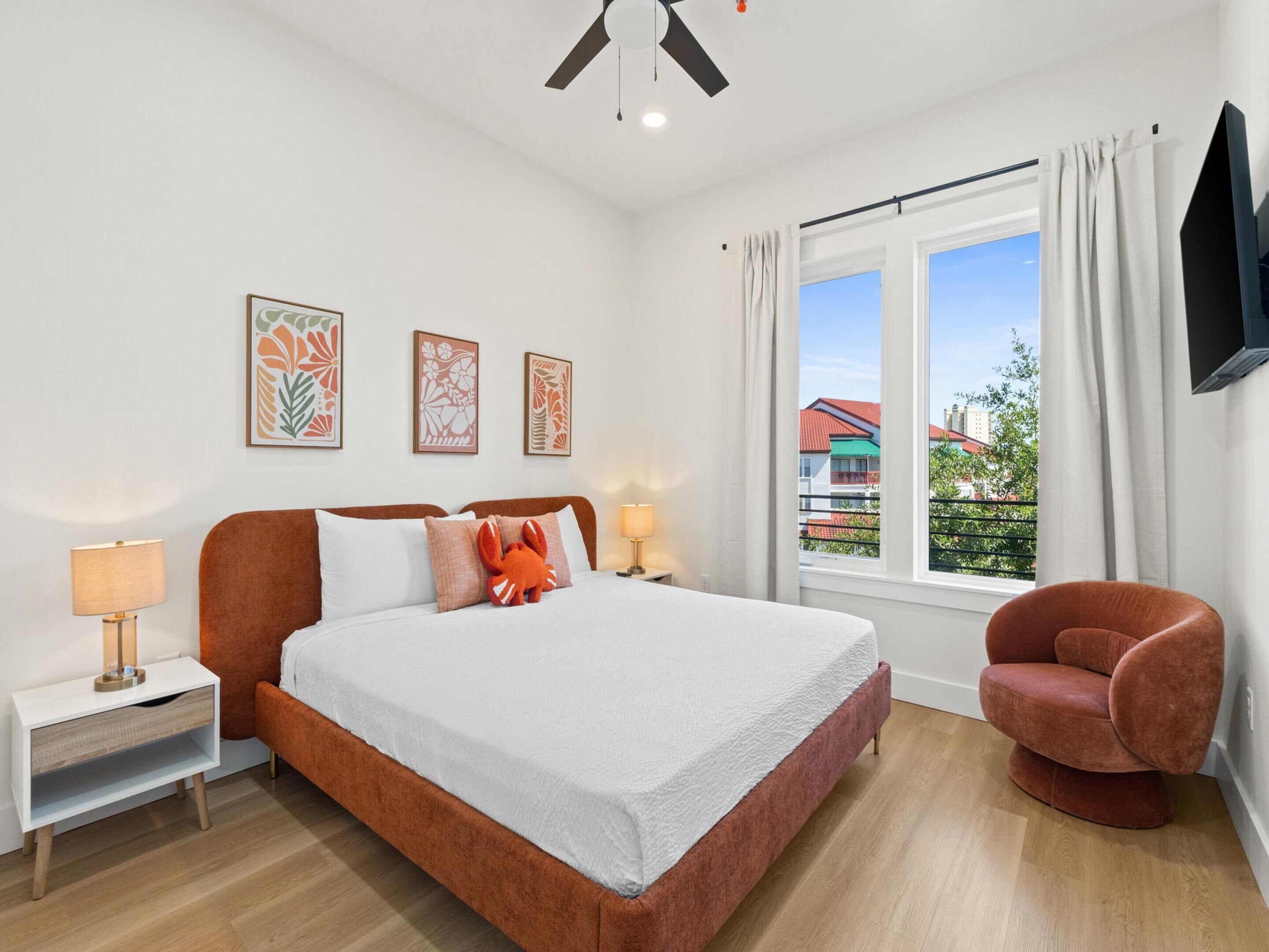 A bedroom with a bed, a nightstand, a chair, a window with a view, and artwork on the wall.