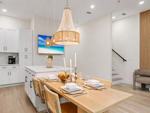 Una cocina moderna con armarios blancos, una mesa de comedor de madera para cuatro personas, una lámpara colgante y un televisor que muestra una escena de playa.