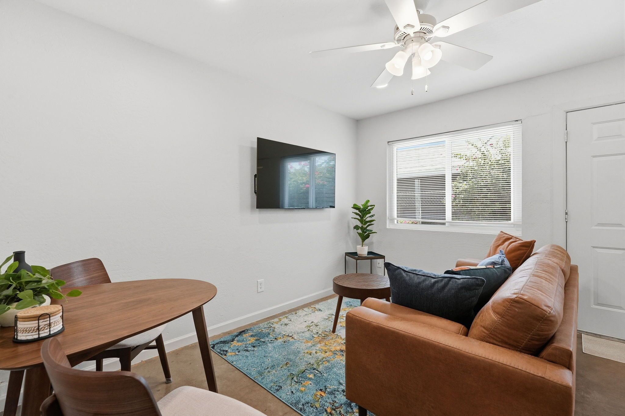 A modern living room with a dining table, a sofa, a ceiling fan, a TV mounted on the wall, and a window with blinds.