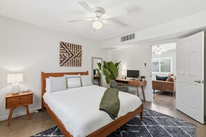 Ein Schlafzimmer mit Bett, Nachttisch, Lampe, Schreibtisch, Stuhl und einer großen Pflanze.
