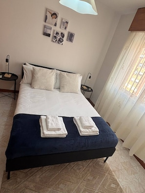 Ein ordentlich bezogenes Bett mit Kissen, zwei Nachttische, eine Wand mit gerahmten Bildern und ein Fenster mit Vorhängen.