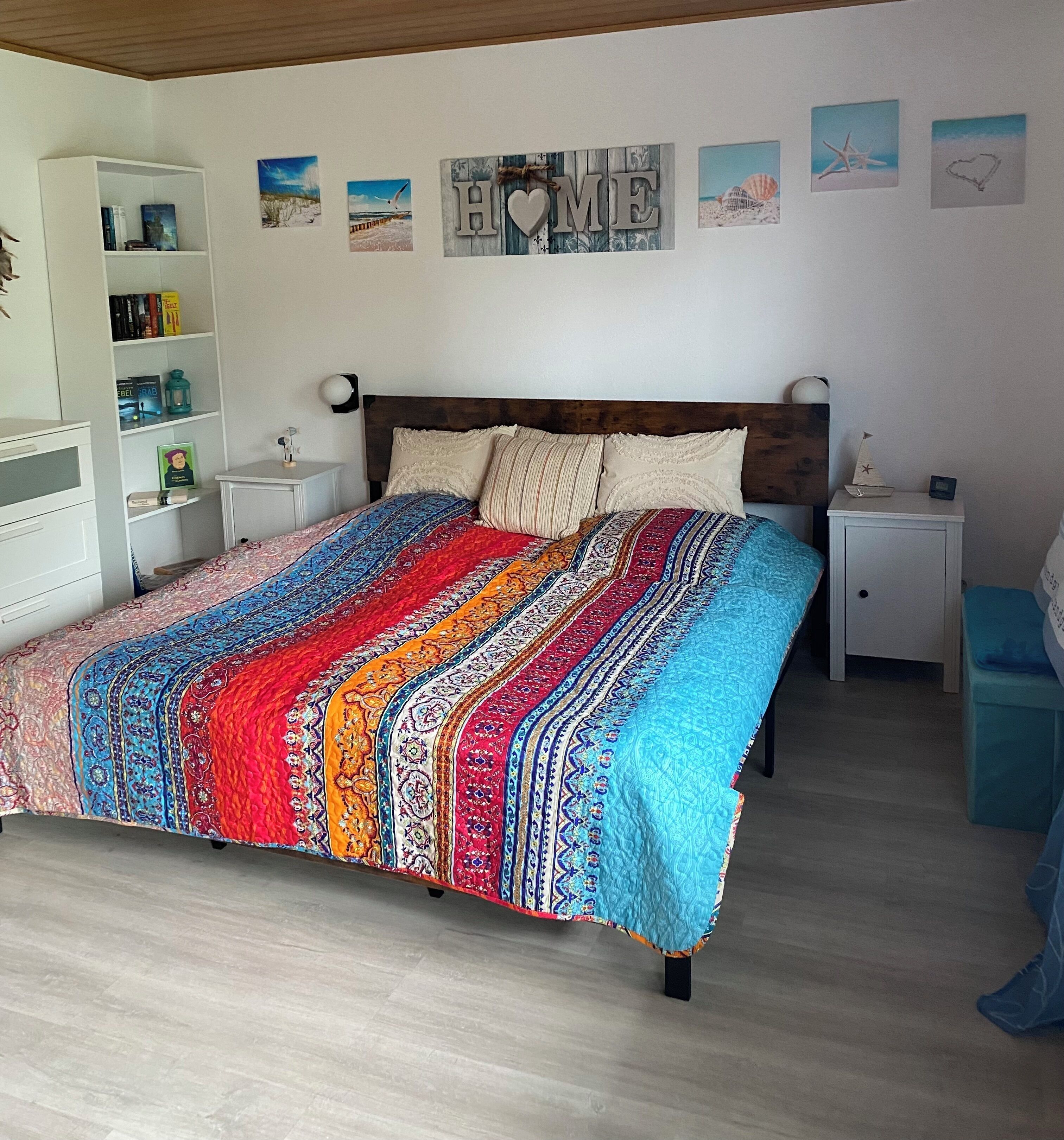 A bedroom with a wooden headboard, a colorful bedspread, a bookshelf, and wall art.