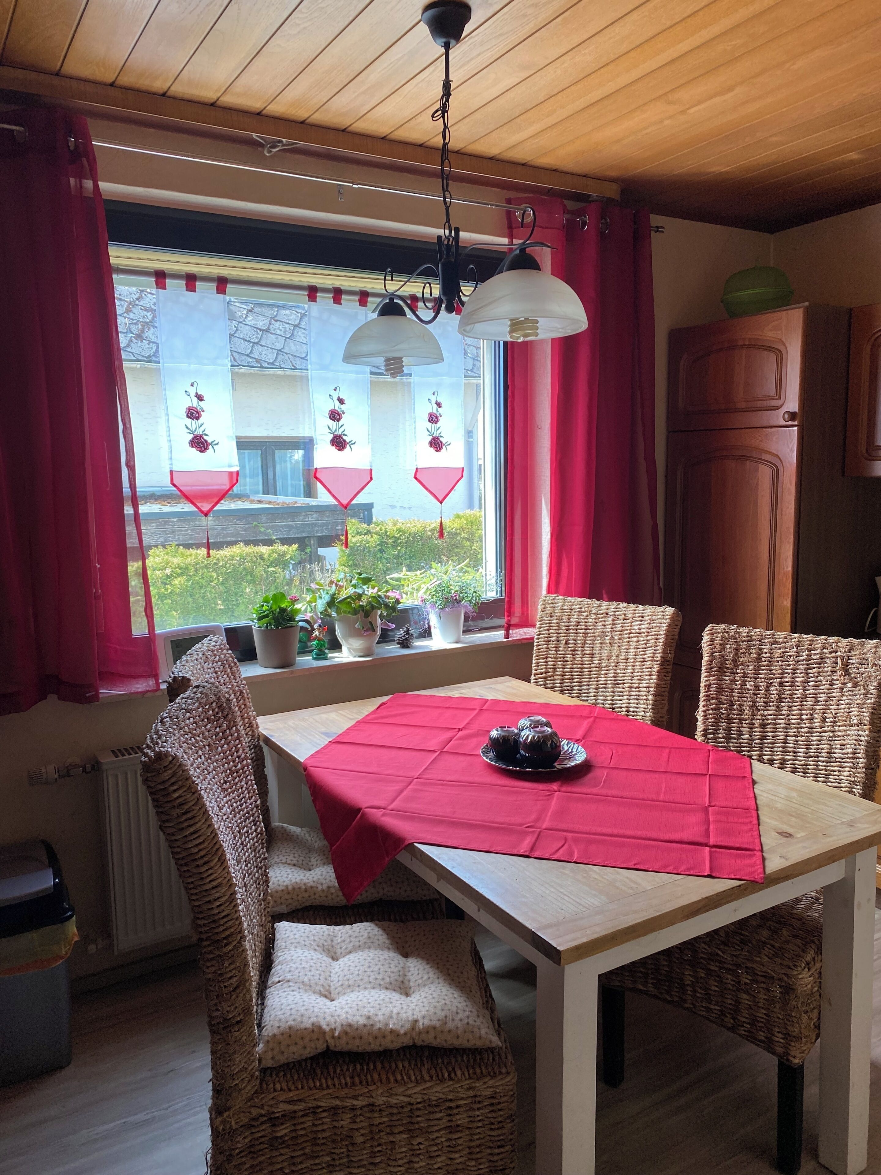 A dining area with a table set for four, wicker chairs, a red tablecloth, and a view of the outside through a window with red curtains.