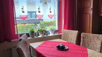 A dining area with a table set for four, wicker chairs, a red tablecloth, and a view of the outside through a window with red curtains.