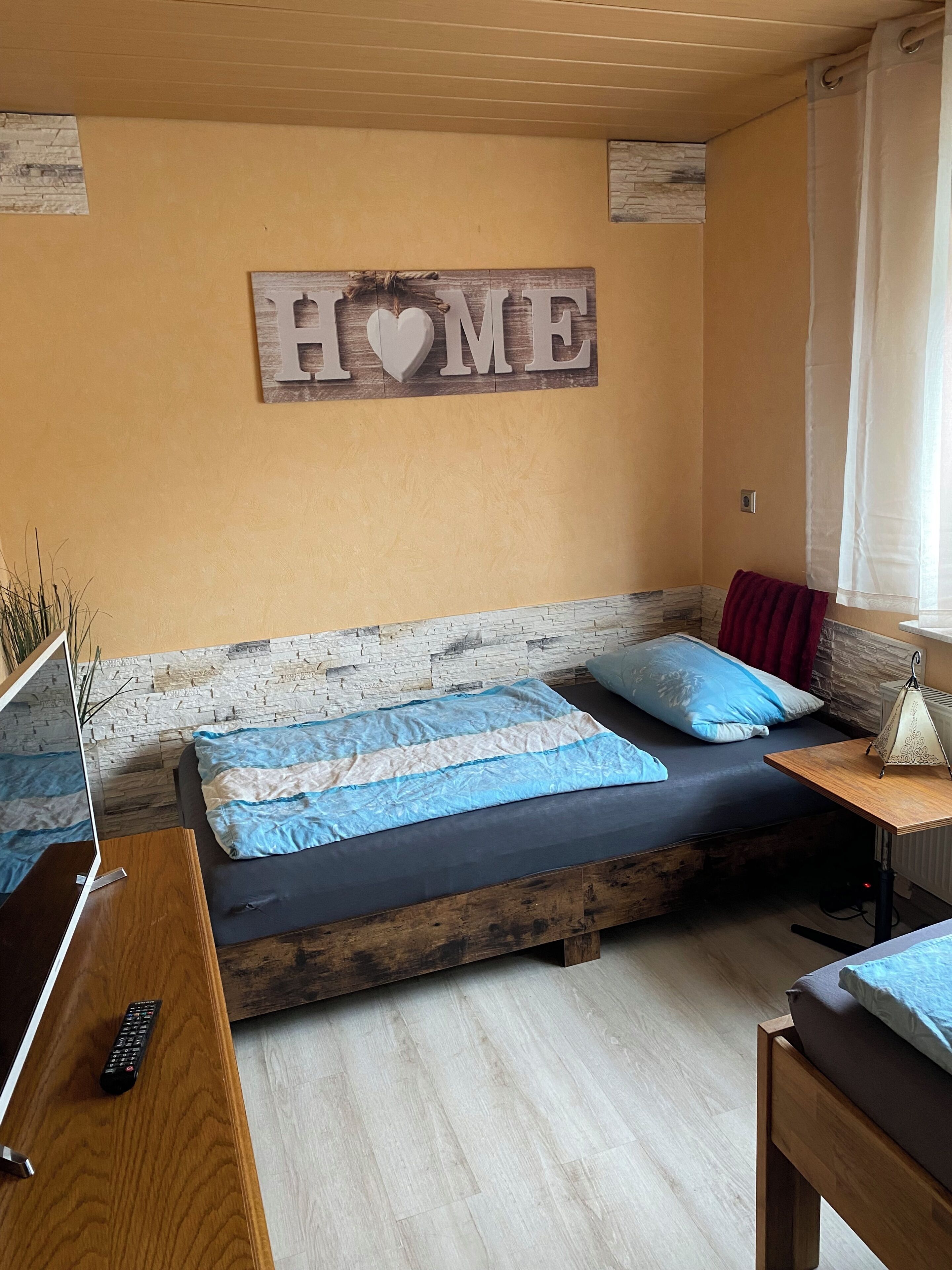 Ein Schlafzimmer mit einem Bett, einem Nachttisch, einer Lampe, einem an der Wand befestigten Schild und einem Fenster mit Vorhängen.