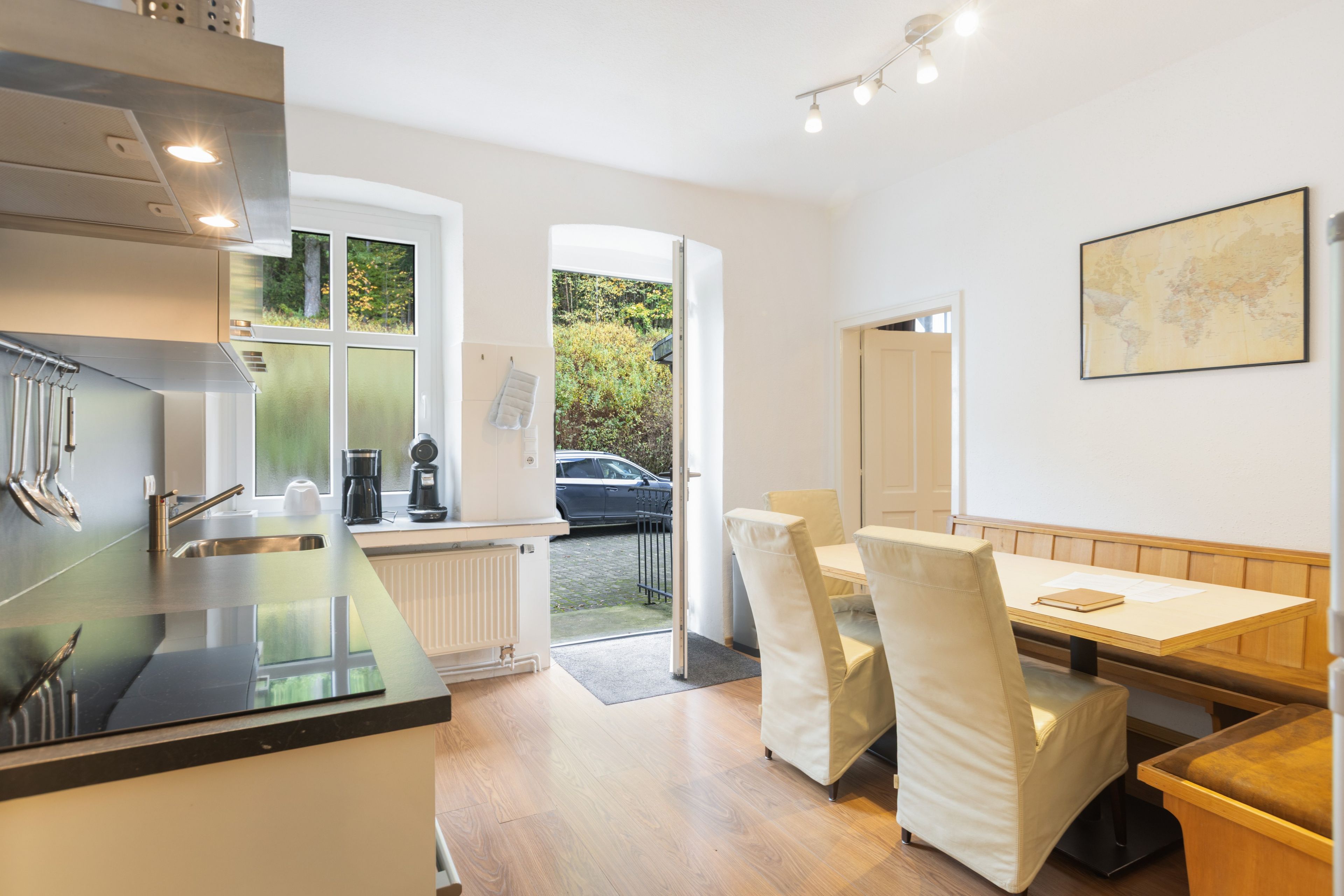 A modern kitchen with a built-in oven, a coffee machine, and a sink. There is a dining area with a table and chairs, and a view of a car parked outside.