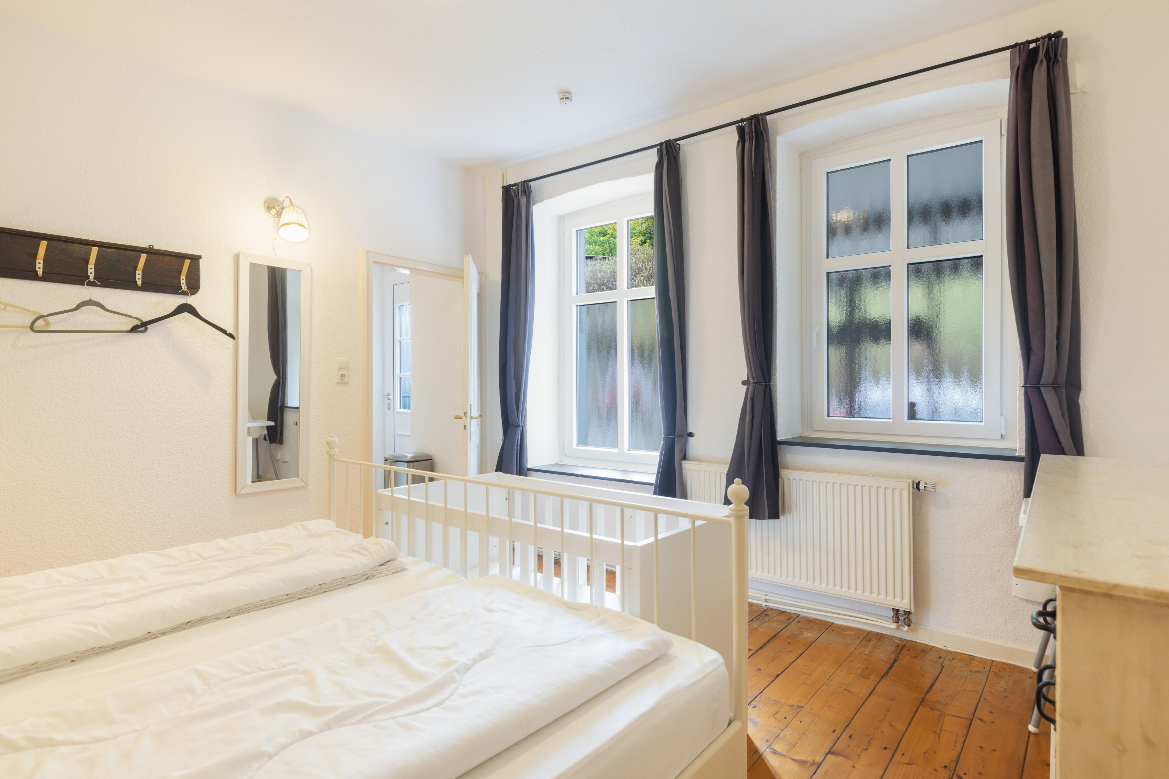 Un dormitorio con cama blanca, ventana con cortinas, radiador y perchero con ganchos.