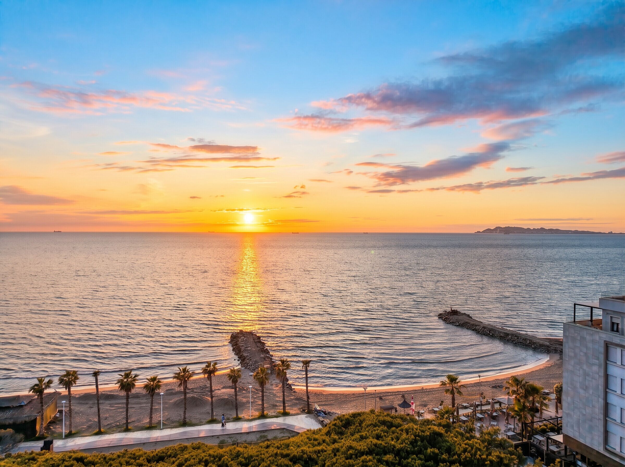 Een kustzonsondergang met een rotsachtige zeewering, palmbomen en een strand.