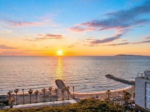 Een kustzonsondergang met een rotsachtige zeewering, palmbomen en een strand.