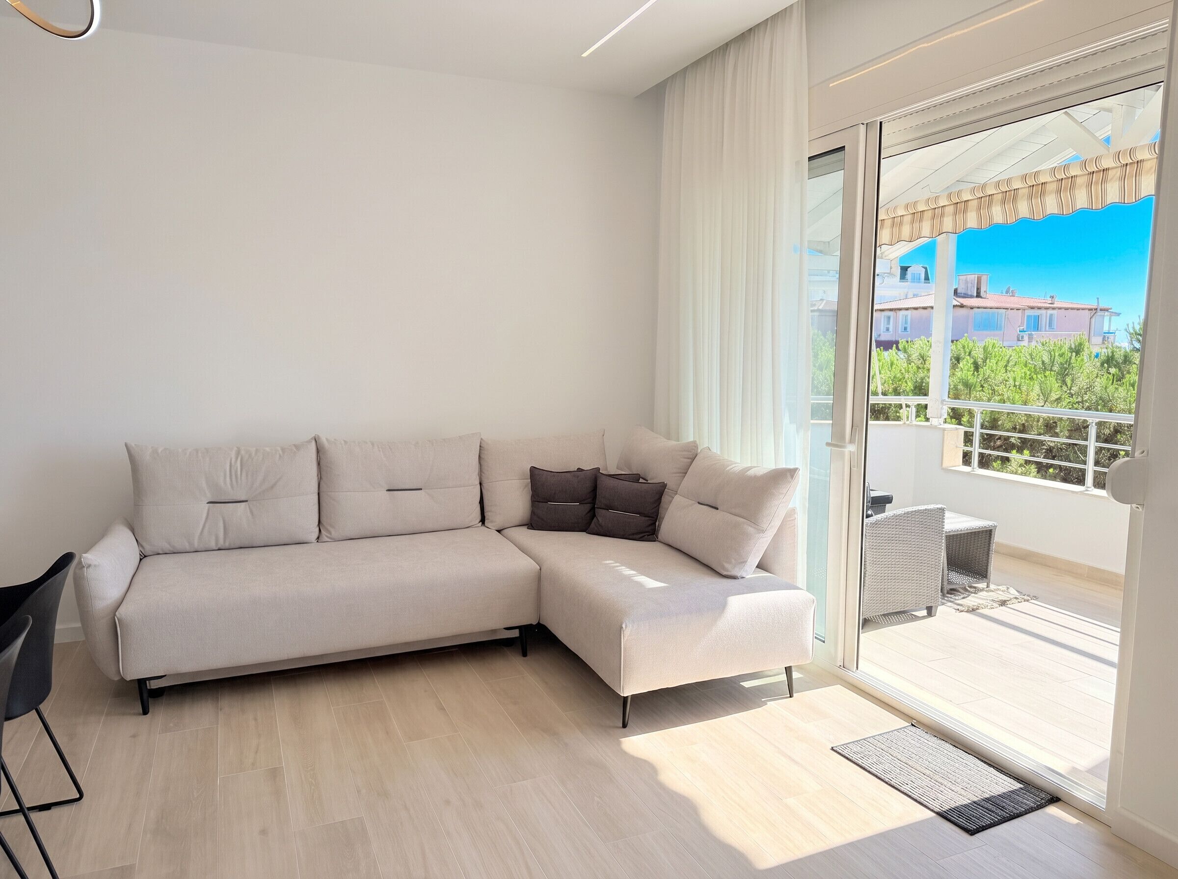 Un salon moderne avec un canapé d’angle beige, un petit fauteuil noir et une porte vitrée donnant sur un balcon d’où l’on peut admirer les bâtiments et la verdure environnante.