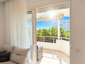A balcony with a view of greenery and buildings, a sofa with cushions, and a sliding glass door. - Sea Breeze Apartment with Balcony (Durres)