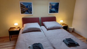 Ein Doppelbett mit zwei Kissen, ein Nachttisch mit Lampe, ein Mülleimer und zwei gerahmte Bilder an der Wand.