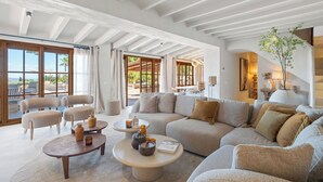 A spacious living area with a large sofa, two armchairs, and a coffee table. There are large windows with curtains, a view of the outdoors, and a staircase leading to an upper level. - Vacation home Prana by Interhome (Moraira)