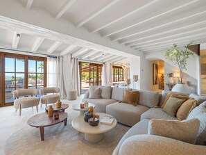 A spacious living area with a large sofa, two armchairs, and a coffee table. There are large windows with curtains, a view of the outdoors, and a staircase leading to an upper level. - Vacation home Prana by Interhome (Moraira)