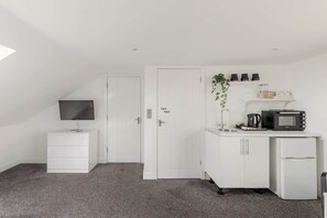 Una habitación pequeña y blanca con kitchenette, televisión de pantalla plana, un armario blanco y una puerta con picaporte.