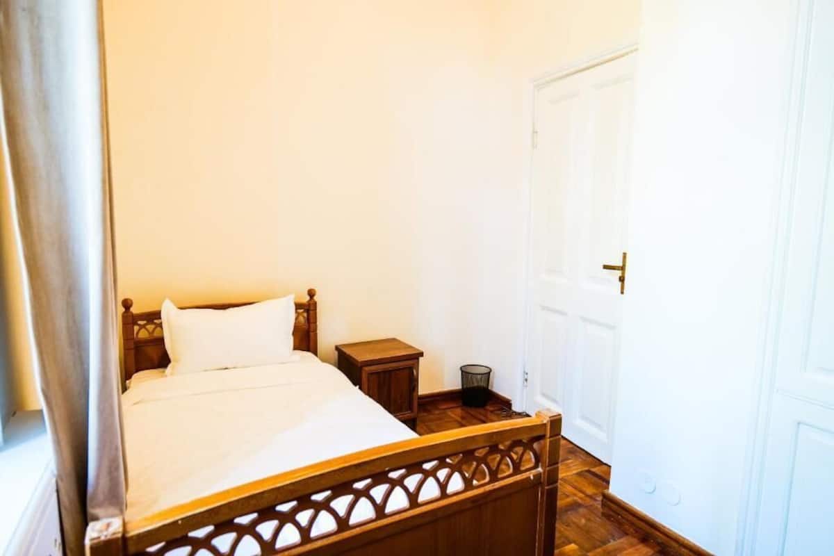 Una cama individual con cabecera de madera, una mesita de noche pequeña y una puerta blanca en una habitación con paredes blancas y suelo de madera.