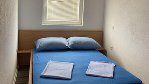 A small, single-bed room with a wooden headboard, a window with blinds, and a blue bedspread. - Apartment Vir 1 (Vir)