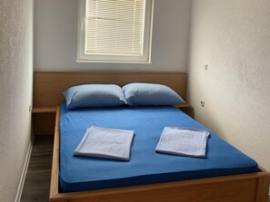 Ein kleines Zimmer mit einem Einzelbett, einem Holzkopfteil, einem Fenster mit Jalousien und einer blauen Bettdecke.