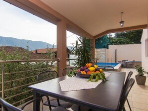 A covered outdoor dining area with a table set for two, a bowl of fruit, and a view of mountains in the background. - Holiday Home Vele With Pool (Vrgorac)