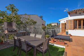 Eine Terrasse mit Grill, Esstisch und Stühlen, umgeben von Kübelpflanzen und einem Zaun.