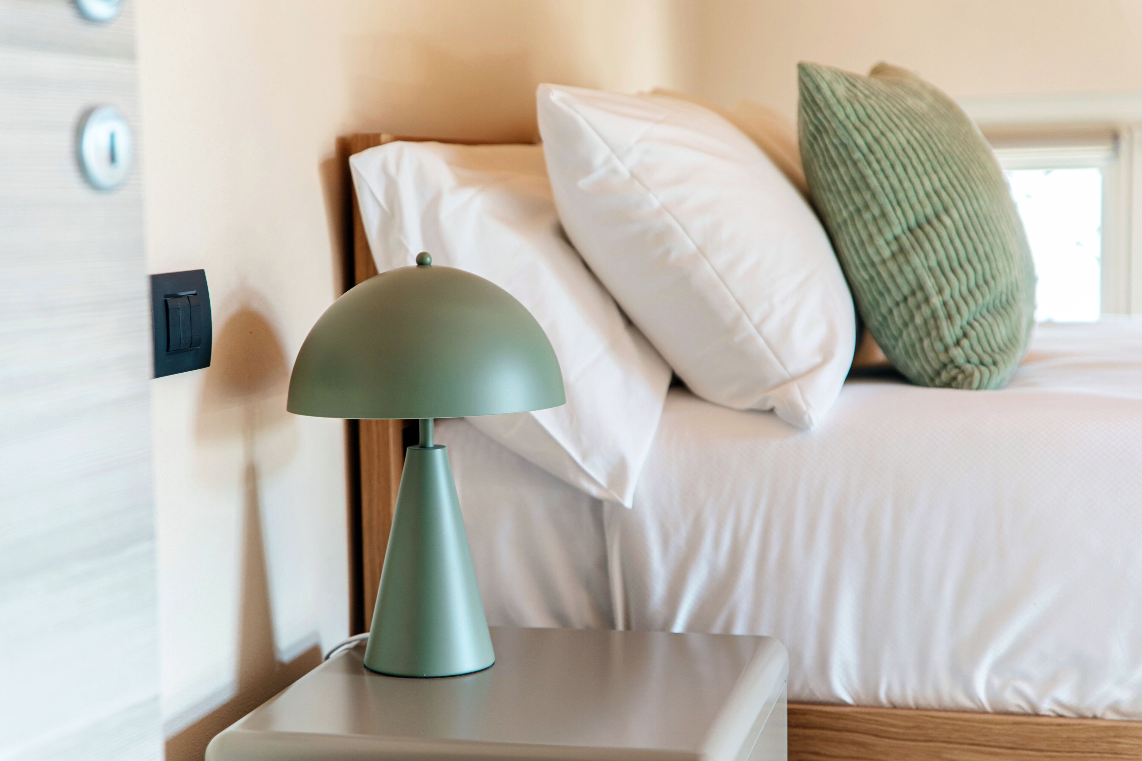 A bed with white bedding, a green lamp on a bedside table, and a window with blinds.