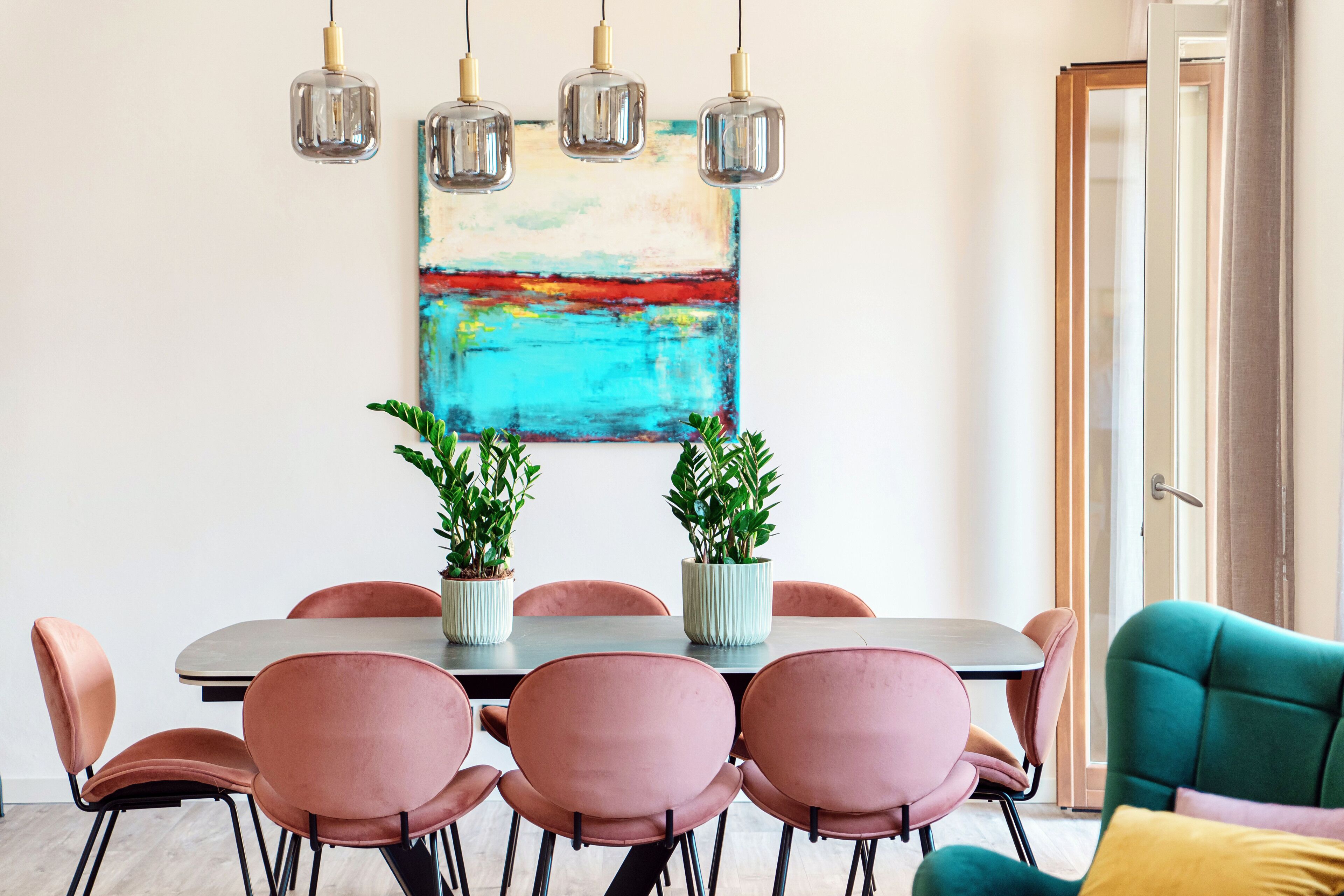 A dining area with a table, chairs, hanging lights, and a colorful abstract painting.