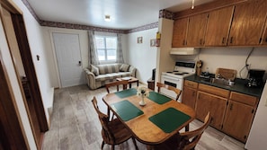 A compact kitchen with wooden cabinets, a dining table with chairs, a sofa, and a window with curtains.