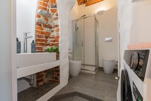 Une salle de bain avec un mur d’accent en briques, une douche en verre, un lavabo blanc et des toilettes.