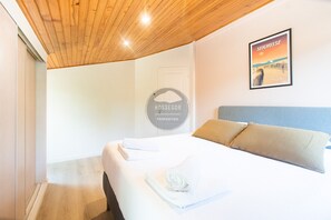 A hotel room with a wooden ceiling, a bed with white linens, a framed picture on the wall, and a sign with 'HOSSEGOR PROPERTIES' on the wall. - Villa Igelak - Hossegor - Center - Lake - Pool (Soorts-Hossegor)
