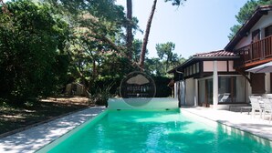 En swimmingpool med et skilt der viser 'HOSSEGOR PROPERTIES', omgivet af træer og en bygning med balkon.