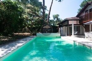 A swimming pool with a sign reading 'HOSSEGOR PROPERTIES' surrounded by trees and a building with a balcony. - Villa Igelak - Hossegor - Center - Lake - Pool (Soorts-Hossegor)