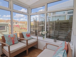 A sunlit room with wicker furniture, large windows, and a view of a wooden fence and buildings. - 2 Lyme Mews (Seaton)