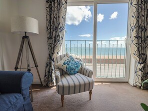 A room with a striped armchair, a lamp, and a view of the beach through a balcony. - 2 Lyme Mews (Seaton)