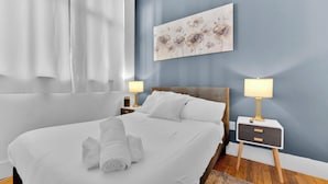 A neatly made bed with white linens, a rolled towel, two bedside tables with lamps, and a floral painting on the wall. - Apartmint at Arch St Philadelphia (Philadelphia)